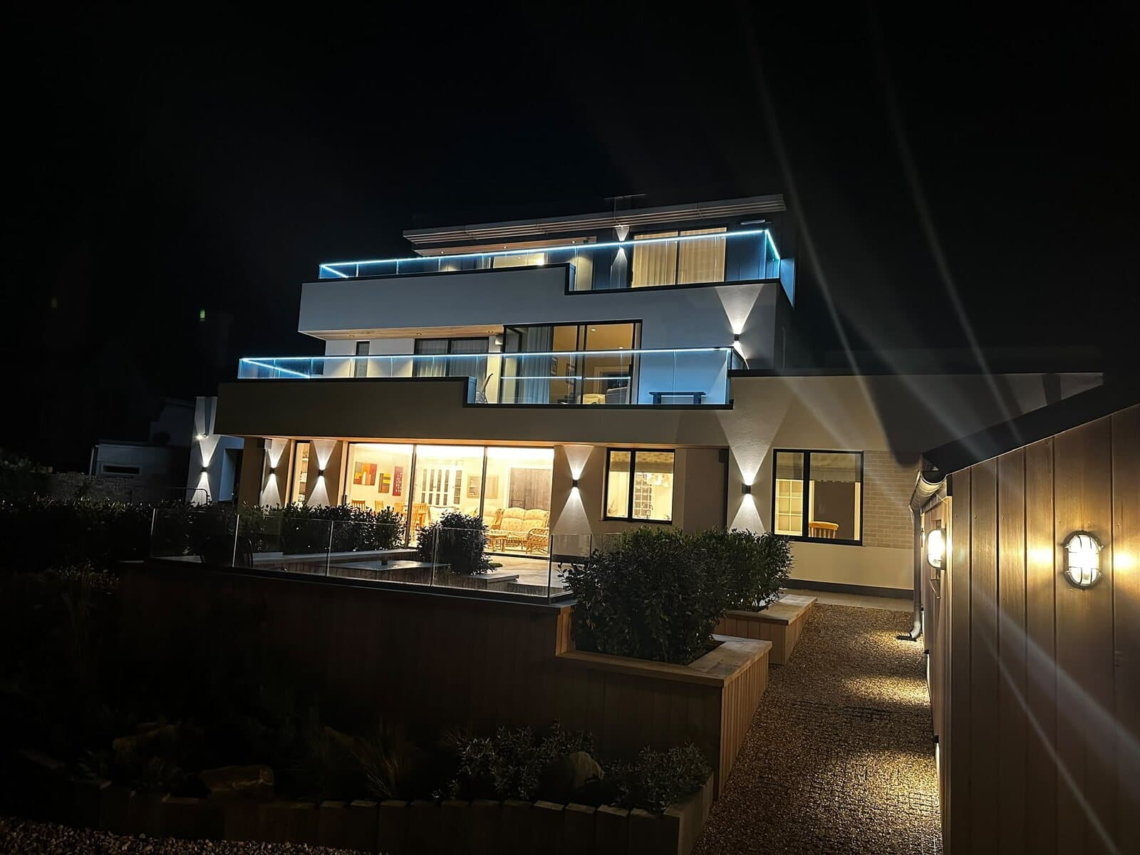 Three-storey new build seen from the garden at night, with lit glass balconies, wall lights and a lit garden path